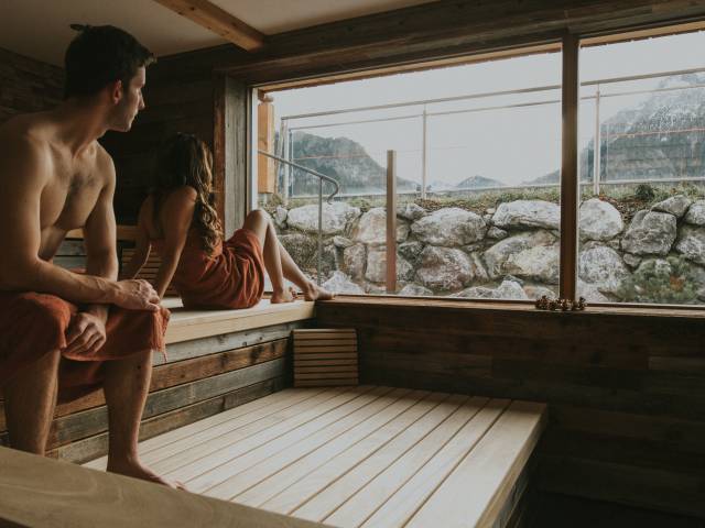 Personen sitzen in Panoramasauna mit Bergblick